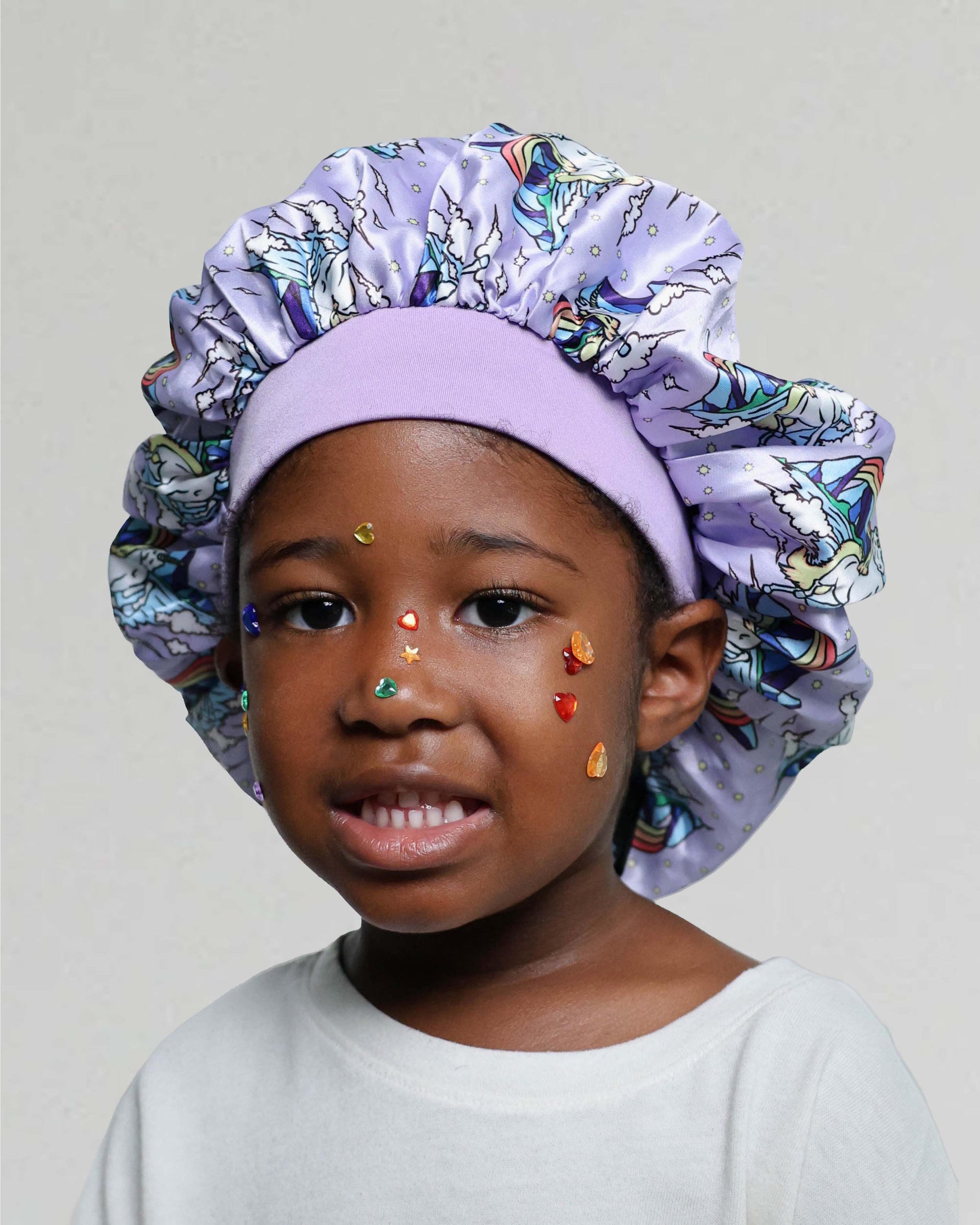 A young child wearing a lavender satin bonnet with a colorful unicorn print and a matching lavender headband, against a plain light gray background.