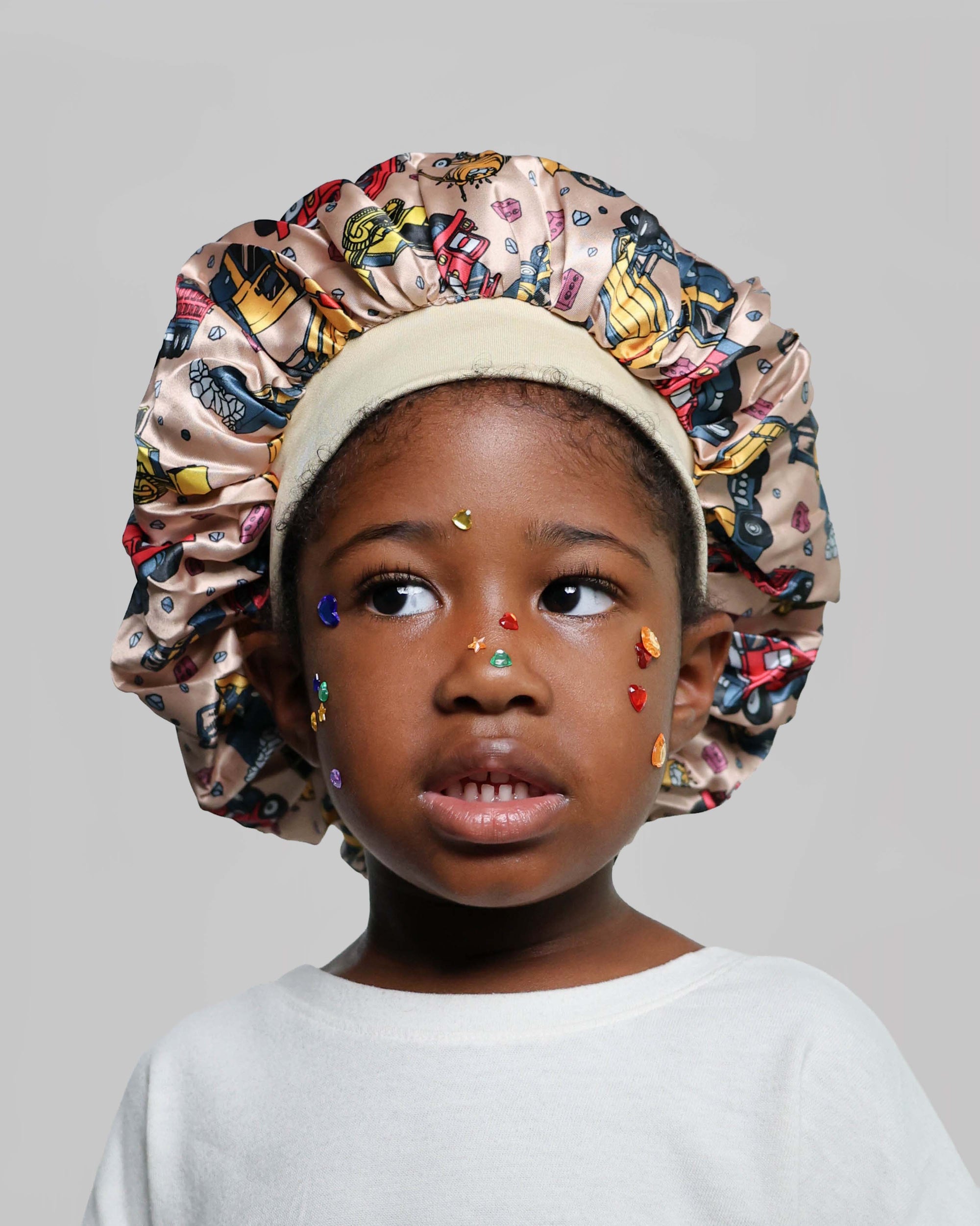 A young child wearing a beige satin bonnet with a colorful construction truck print and small multicolored face gems, photographed against a plain light gray background.