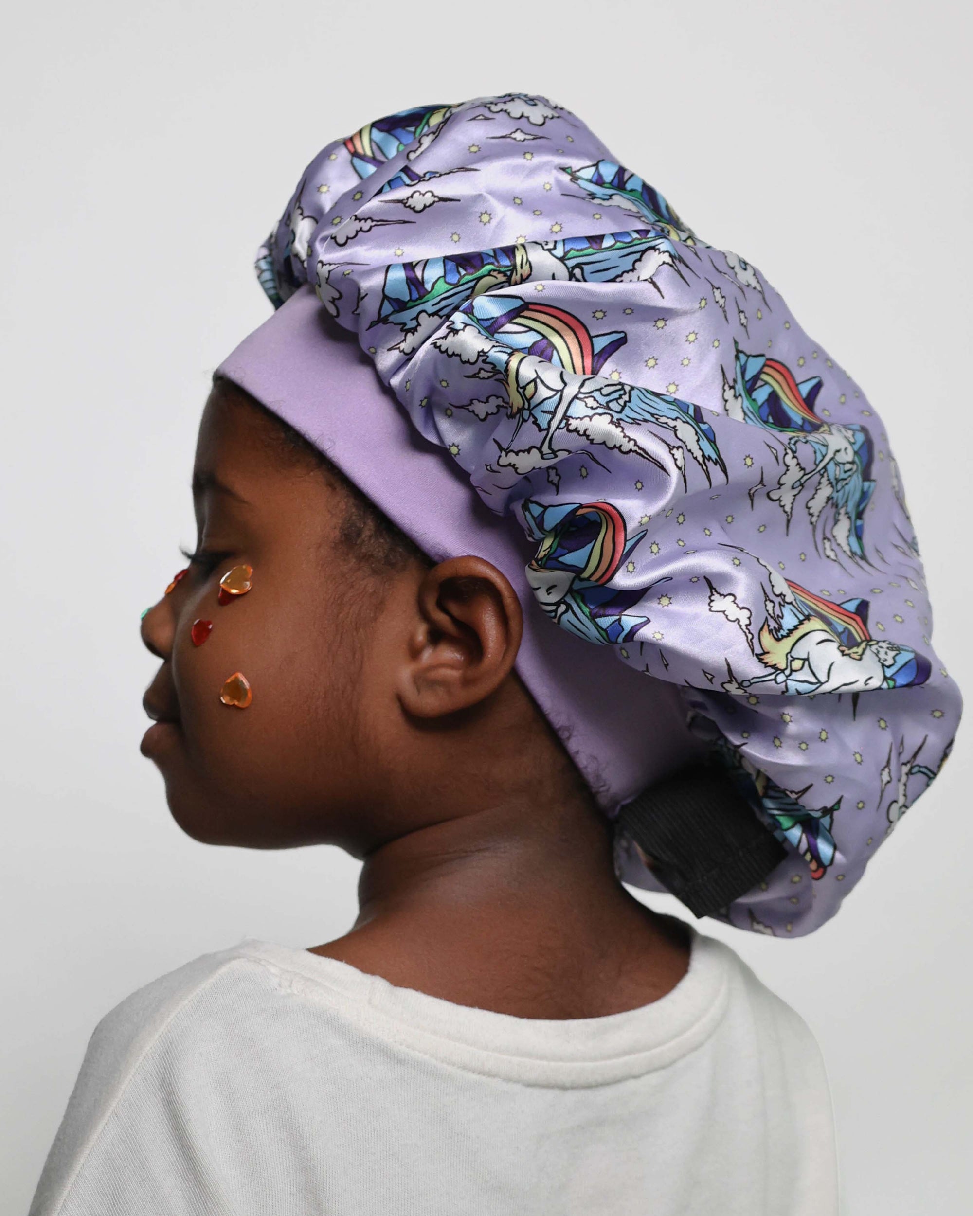 A child shown in profile wearing a lavender satin bonnet with a colorful unicorn print. The bonnet features a solid lavender inner band and a black elastic back closure.