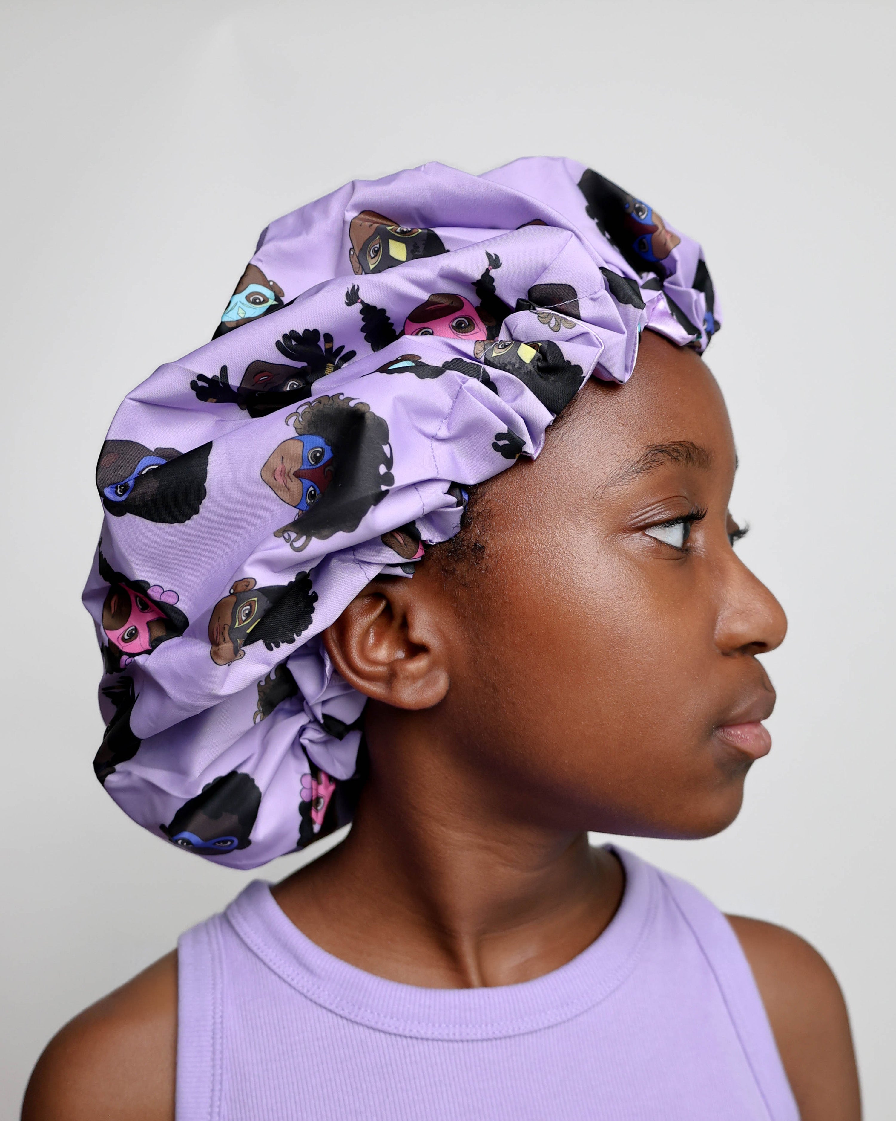 Child wearing a purple shower cap with black and brown superhero's on a plain background