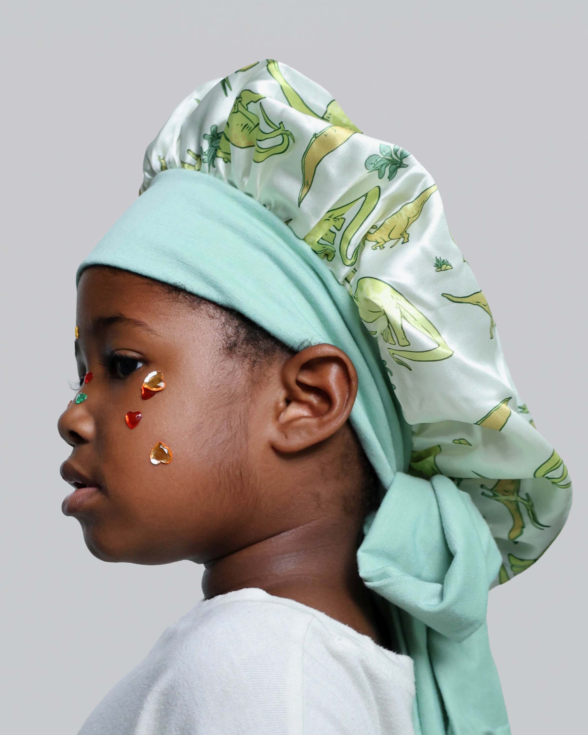 A child in profile wearing a mint green satin bonnet with a light green and yellow botanical print, along with small red and gold face stickers, against a solid gray background.