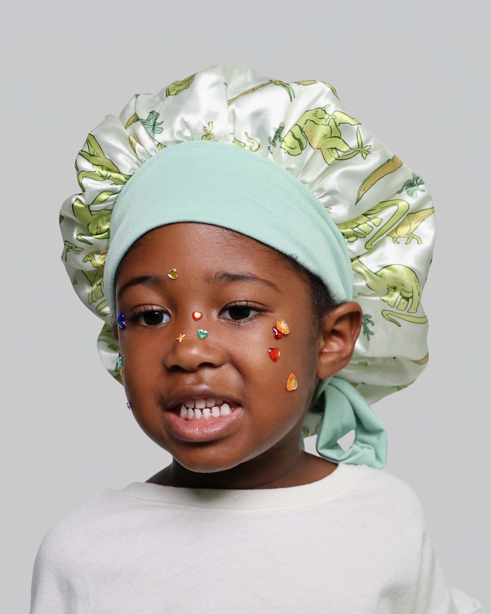 A young child wearing a mint green satin bonnet with a green dinosaur print and a white long-sleeve shirt, against a solid gray background.